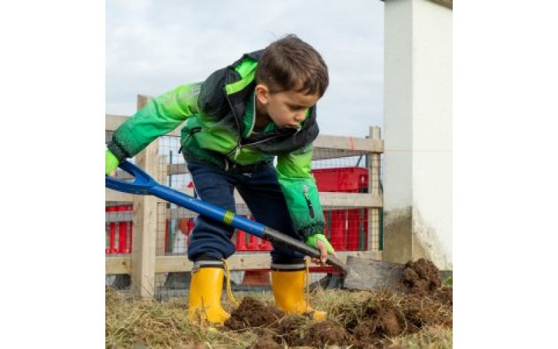 Child in wellingtons digging sand with a large spade