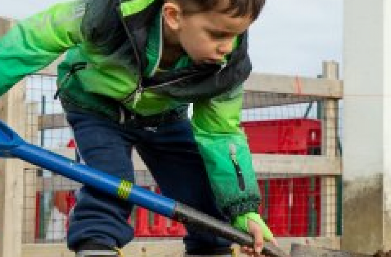 Child in wellingtons digging sand with a large spade