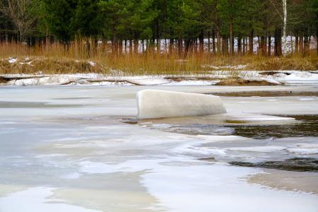 Large lump of ice on a river covered with broken ice, there are some trees in the background on the shore.