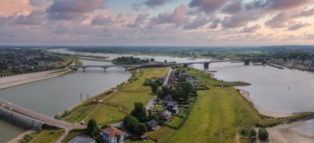 Aerial view of Nijmegen, Netherlands, showcasing the Waal River, De Oversteek, Waalbrug, green fields, residential areas, and scattered clouds in the sky.