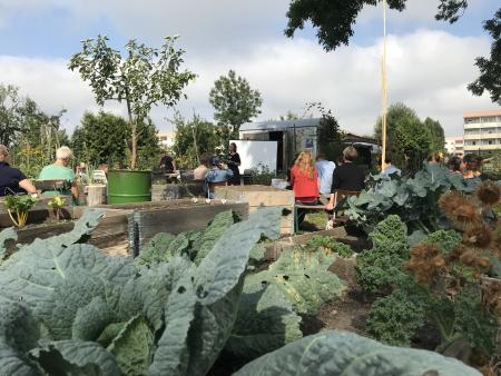 People sitting in a circle in a community garden listening to a speaker who is standing up.